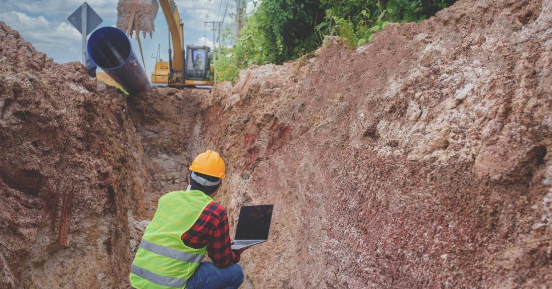 A young Asian engineer is inspecting a large sewer that is buried underground at a construction site. The excavator is empty the water pipe.
